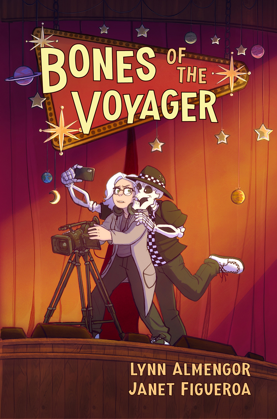 Cover of the graphic novel, “Bones of the Voyager” by Lynn Almengor and Janet Figueroa. A young woman with white hair and glasses stands on a stage decorated with a red curtain and stars hanging from the ceiling by strings. She’s trying to use a video camera while a skeleton wearing a black-and-white checkered ska getup interrupts from behind her to take a selfie.
